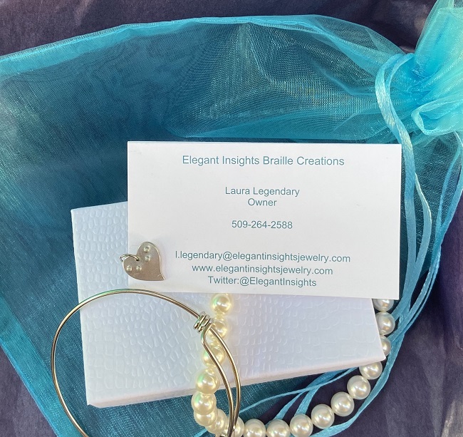 Mixed composition photo representative of the Sparkle Dots subscription. A small white box and a teal jewelry pouch rests on purple tissue paper at front left. A strand of pearls is draped from the box, along with a wire bracelet. At top is an Elegant Insights business card next to a heart charm embossed with E I.