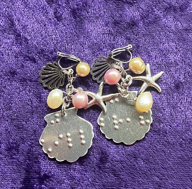 Photo of the Seaside Serenity Dangle Earrings in the clip style for non-pierced ears, braille-embossed with "just breathe," on one earring, and "just chill," on the other.