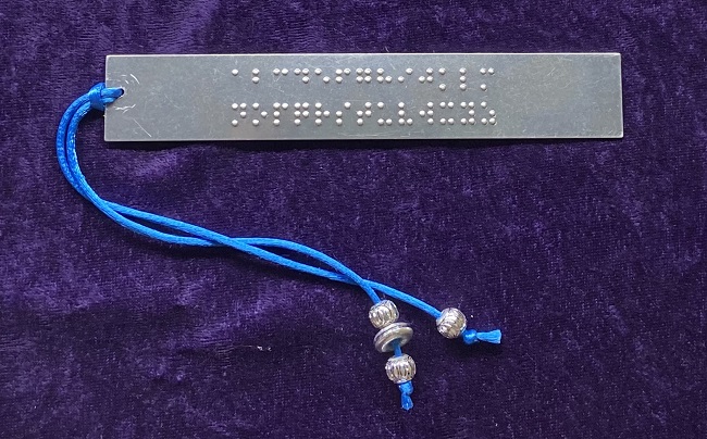 Photo of aluminum bookmark, embossed with the braille alphabet in two rows. A blue silk cord decorated with silvery beads drapes from right to left.