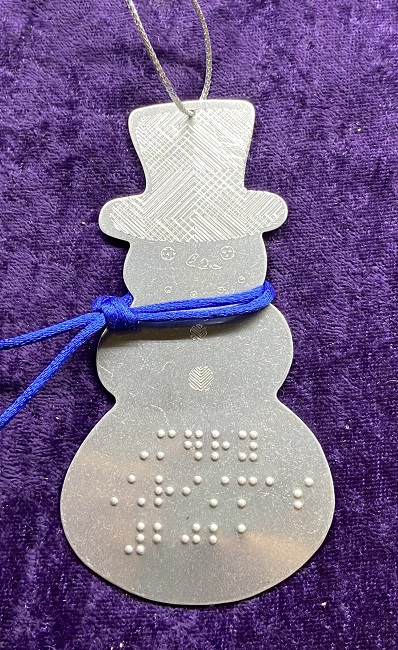 Photo of the Snowman Holiday Ornament, braille embossed with Merry Christmas 2021. He has an engraved face and buttons, a textured,patterned hat, and he is wearing a blue silk cord scarf.
